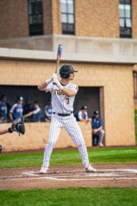 Baseball player in a white striped uniform stands ready to bat on the field with teammates in the background.