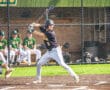 Young baseball player in uniform swings bat during a game, spectators in green jerseys visible in the background.