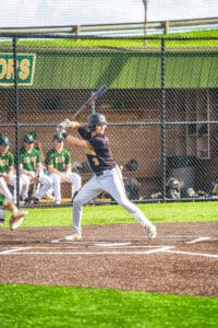 Young baseball player in uniform swings bat during a game, spectators in green jerseys visible in the background.