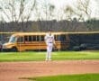High school baseball player in a striped uniform stands on the field with an orange school bus in the background.