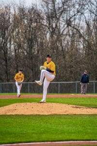 Baseball pitcher on the mound in a yellow team jersey, preparing to throw during a game, with teammates in the background.