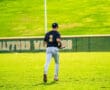 Baseball player in jersey number 3 walks on green field with grass, preparing for the game in bright sunlight.