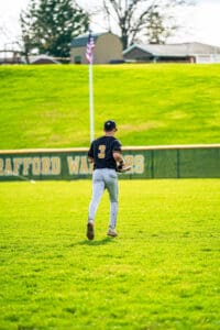 Baseball player in jersey number 3 walks on green field with grass, preparing for the game in bright sunlight.