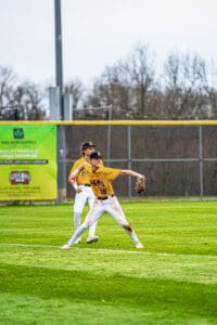 Baseball players in yellow jerseys fielding a ball at a sunny outdoor game, with a green advertisement in the background.