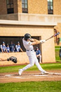 Baseball player in striped uniform swinging bat at home plate, with teammates in the background. Bright sunny day.