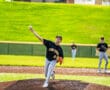 High school baseball pitcher throwing a ball from the mound, teammates in background on a sunny day.