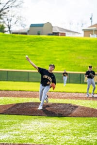 High school baseball pitcher throwing a ball from the mound, teammates in background on a sunny day.