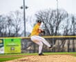 Young baseball pitcher on the mound, preparing to throw, with a grassy field and bare trees in the background.