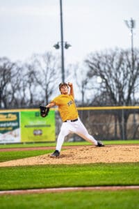 Young pitcher in bright yellow uniform throwing a baseball on a well-maintained field, with trees and lights in the background.