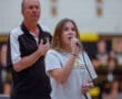 Student singing at a school event, with coach in the background and cheerleaders in the distance, creating a lively atmosphere.
