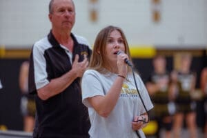 Student singing at a school event, with coach in the background and cheerleaders in the distance, creating a lively atmosphere.
