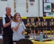 Girl singing national anthem with microphone, while coach stands behind her and basketball team lines up in background.