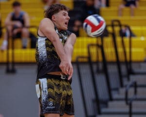 High school volleyball player prepares to pass a ball during a match in a gym with spectators in the background.