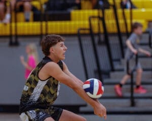 Teenage boy preparing to serve a volleyball during a game in a gymnasium, with spectators in the background.
