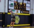 Teenage volleyball player serves during a match, with scoreboard showing 14-12 score in a gym setting.