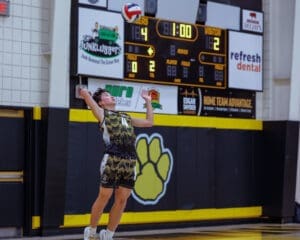 Teenage volleyball player serves during a match, with scoreboard showing 14-12 score in a gym setting.