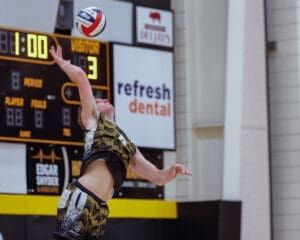 A volleyball player prepares to serve, captured in action during a competitive match with a scoreboard in the background.