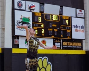 Volleyball player serving during a match, score: Tigers 18, Visitor 4, with 1 minute left in the period.