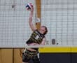 A young male volleyball player jumps to serve the ball in a gym with a beige and yellow backdrop.