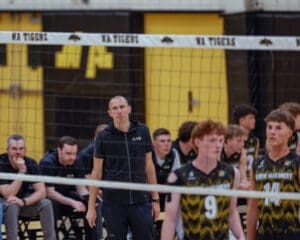 Volleyball coach watches the game intently, surrounded by players and assistants on the sidelines at a high school match.