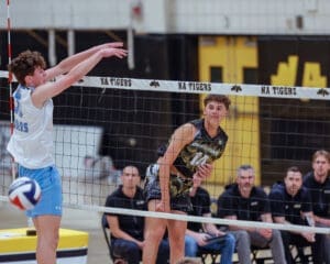 High school boys' volleyball match: player spikes ball over the net while teammate prepares for defense in a competitive atmosphere.