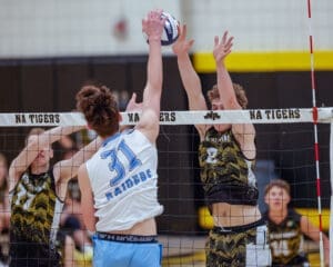 High school volleyball players jump to block an opponent's spike at a competitive match. Action-packed scene at the net.