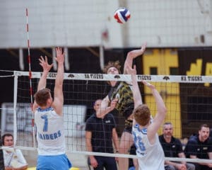 High school volleyball match action, player spiking the ball over the net while defenders jump to block.