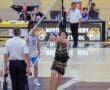 Volleyball match action: player prepares to set the ball as referee watches, with spectators and team names in the background.