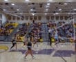 High school volleyball game in action, with players jumping at the net surrounded by an enthusiastic crowd.