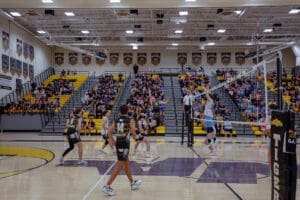 High school volleyball game in action, with players jumping at the net surrounded by an enthusiastic crowd.