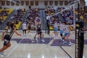 High school boys' volleyball match in action, with players jumping and a packed crowd cheering in the background.