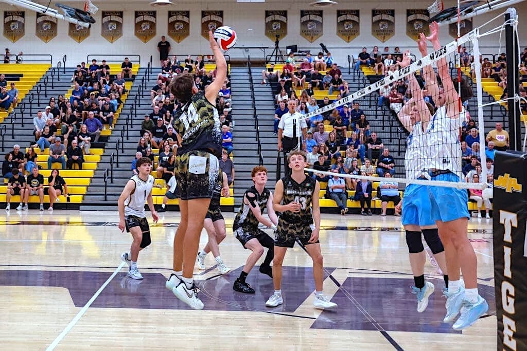 A volleyball player leaps to spike the ball over blockers during an intense match in a packed gymnasium.