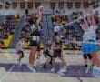 High school volleyball match with players jumping to spike the ball, surrounded by a cheering crowd.