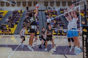 High school volleyball match with players jumping to spike the ball, surrounded by a cheering crowd.