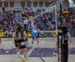 High school volleyball match in action, with players jumping to spike the ball and spectators cheering in the background.