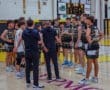 High school basketball team huddling during a game, discussing strategy on the court with a scoreboard in the background.