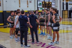 High school basketball team huddling during a game, discussing strategy on the court with a scoreboard in the background.