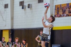 High school volleyball player serves the ball during a match, with teammates in the background, showcasing athleticism.