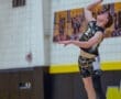 High school athlete jumping to spike a volleyball during a match in a gymnasium, showcasing skill and athleticism.