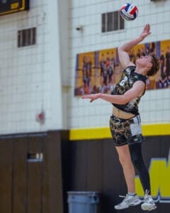 High school athlete jumping to spike a volleyball during a match in a gymnasium, showcasing skill and athleticism.
