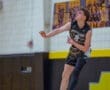 High school volleyball player jumps for a play, wearing a black and gold uniform, in a gym setting.