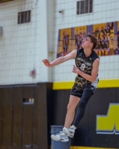 High school volleyball player jumps for a play, wearing a black and gold uniform, in a gym setting.