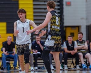 Two young basketball players celebrate on the court after a play, with teammates cheering in the background.