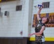 High school volleyball player jumps to serve the ball during a match in a gymnasium with cheering teammates in the background.