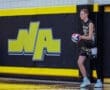 A basketball player stands by the wall, holding a ball in a gym with a vibrant yellow and black backdrop.