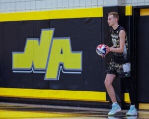 A basketball player stands by the wall, holding a ball in a gym with a vibrant yellow and black backdrop.