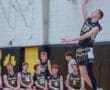 High school volleyball player in mid-air serve, teammates watching on the bench during a competitive match.