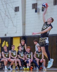 High school volleyball player in mid-air serve, teammates watching on the bench during a competitive match.