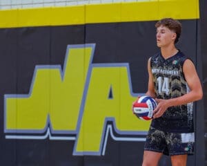 Male volleyball player in North Allegheny uniform preparing to serve, with a vibrant team logo in the background.