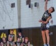 High school volleyball player jumps to serve during a match, with teammates watching in the background.